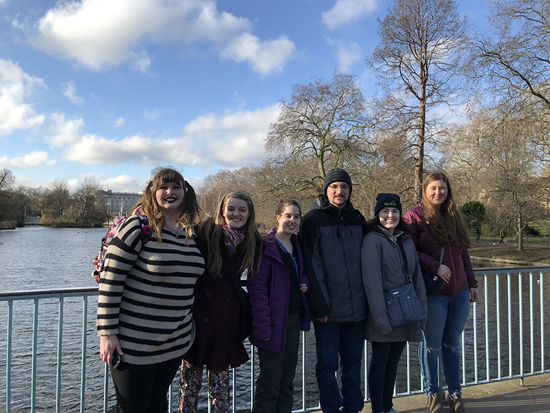 Six students pose for a picture outdoors on a bridge on a sunny day. Their backs are leaning against the railing with a river behind them. Their clothing indicates the weather is chilly.