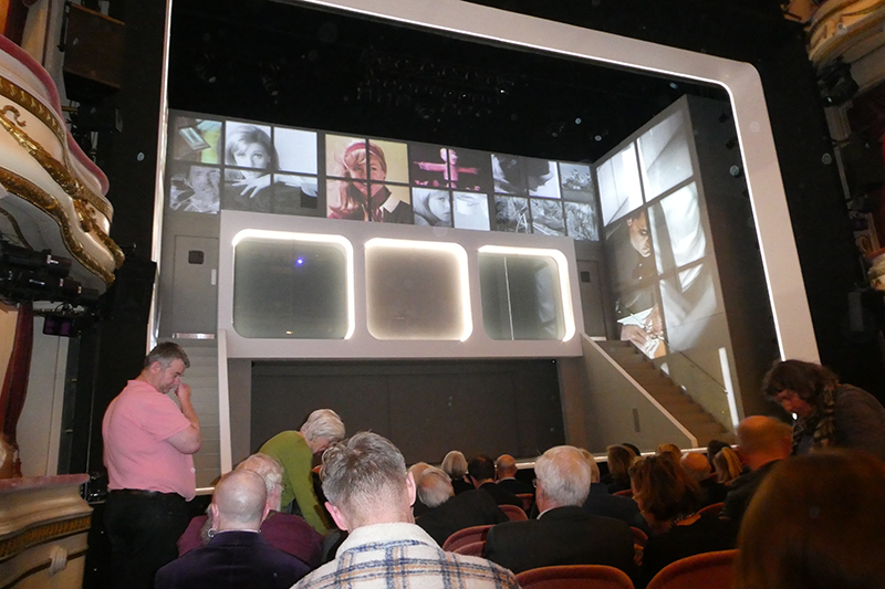 Audience view of stage set, which features images on multiple video screens and staircase leading up the right-hand side.