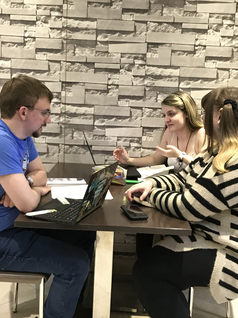 A male student sits alone on the left facing two female students seated side by side on the righ side of a table.