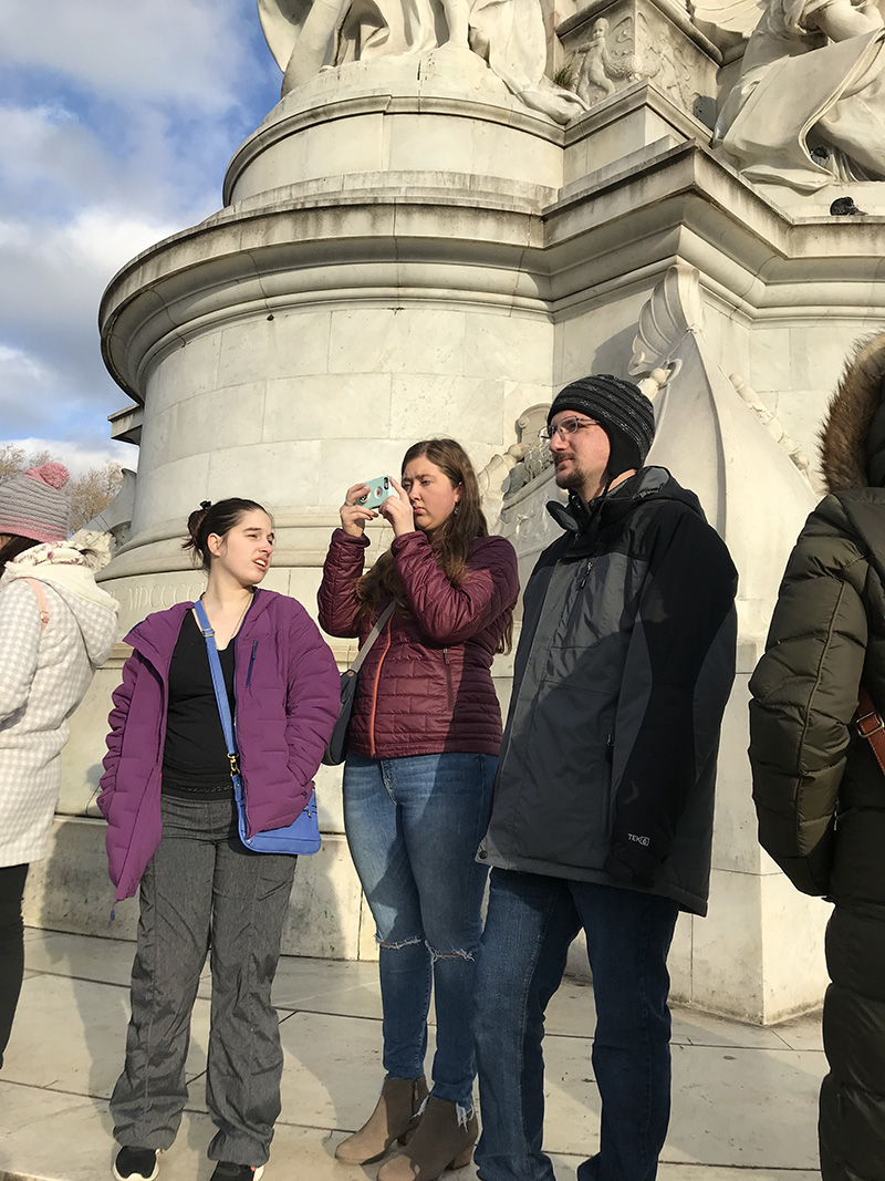A student is holding a camera phone to take a picture of an object located somewhere to the left of the image. A female student on her left is saying something to her. A male student on her right is looking in the same direction that her camera is pointed.
